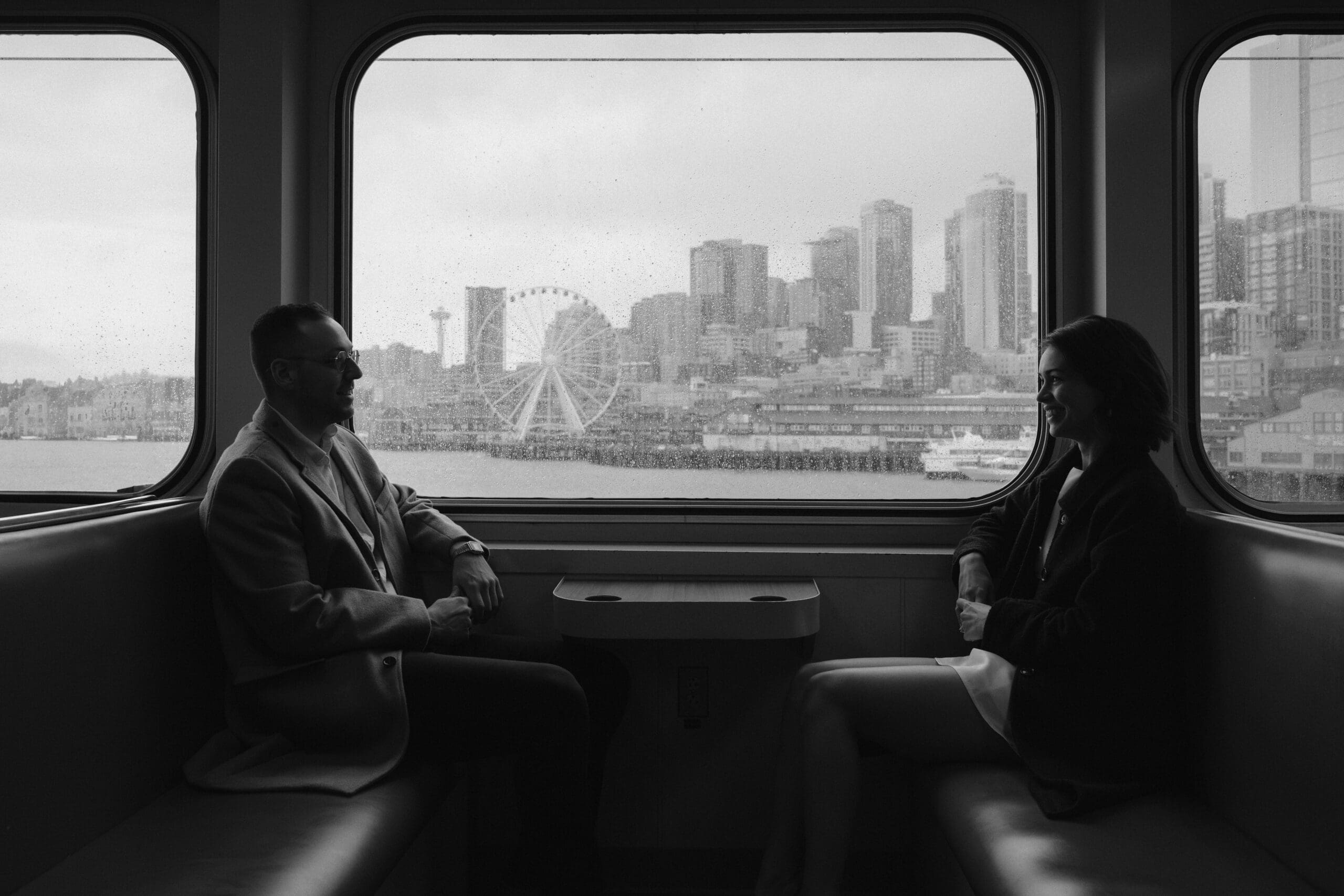 Black and white photo of a couple sitting in a ferry boat booth smiling at each other with the city skyline in the window between them