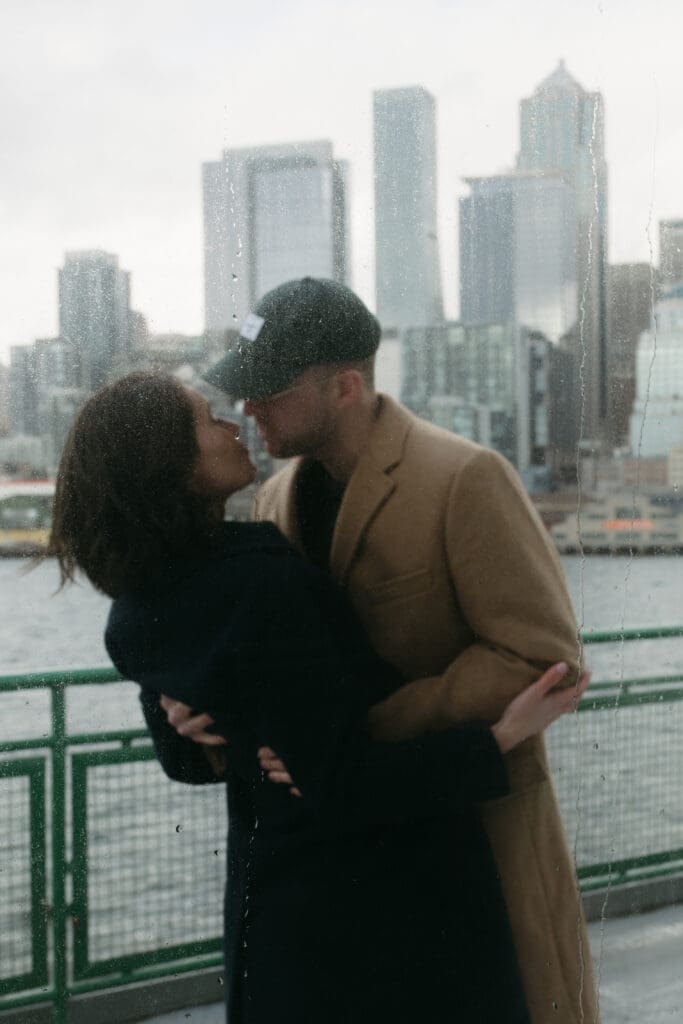 A photo taken through a window of a couple leaning in for a kiss with raindrops rolling down the glass and the Seattle skyline behind the,