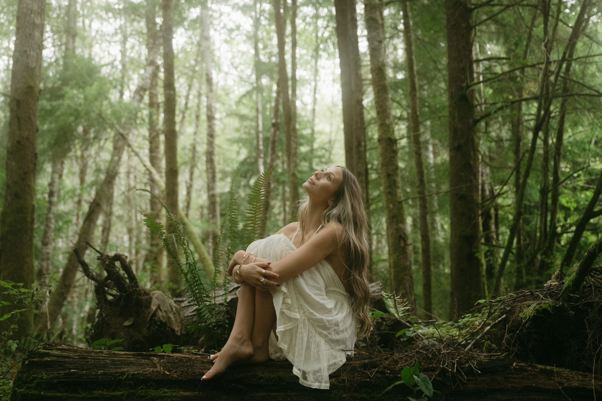 A photo of a girl sitting on a log in the forest gazing upward toward the sky as soft light filters through the trees behind her