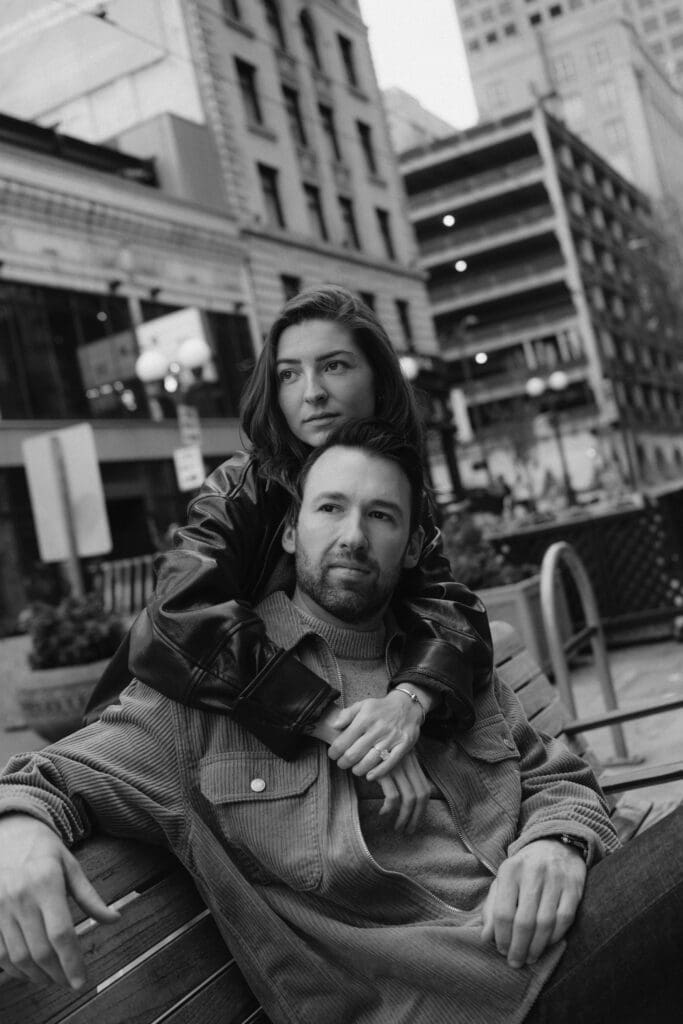 Black and white photo of a man sitting on a bench looking in the distance while a woman wraps her arms around him
