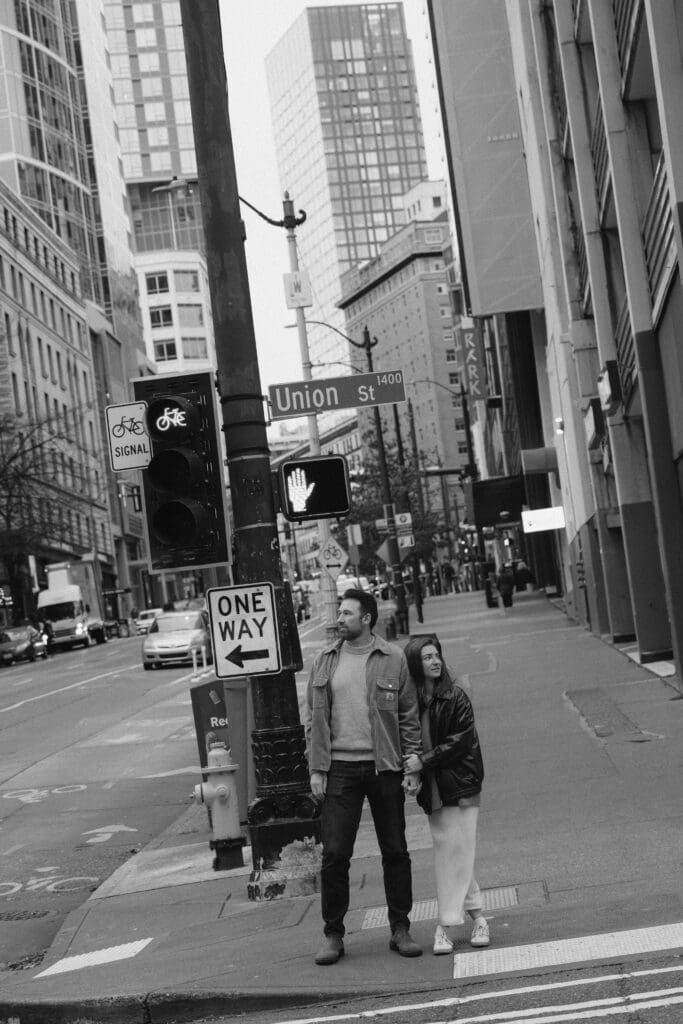 Black and white photo of a woman leaning into a man's arm while waiting to cross the street in the city