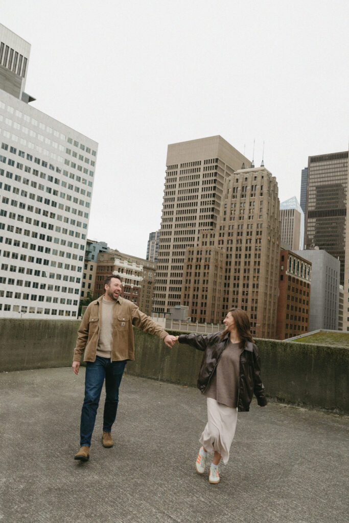 Engaged couple walking toward the camera holding hands with the city skyline in the background