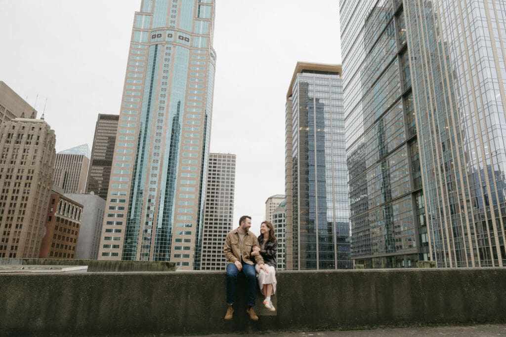 Wide shot of a couple sitting next to each other on a roof with the Seattle skyline in the background