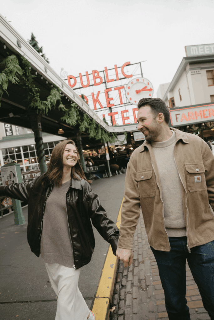 Couple holding hands walking toward camera with Pike Place Market sign behind them